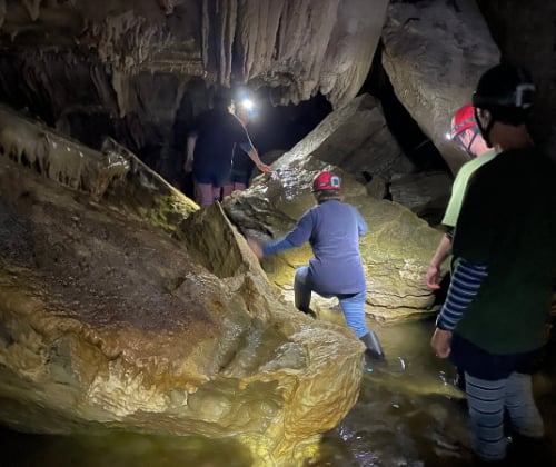 Small tour group navigating rocks and shallow water inside a limestone cave during an underground exploration.