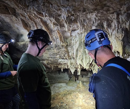 Visitors wearing helmets and headlamps observing stalactites and limestone formations inside a cave.