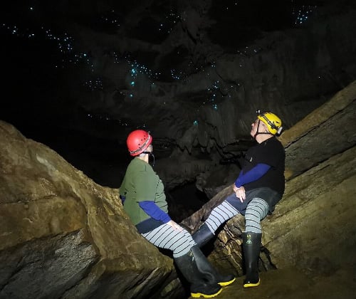 Two visitors standing on a rock ledge beneath glowworms, highlighting the cave’s height and glowing ceiling.