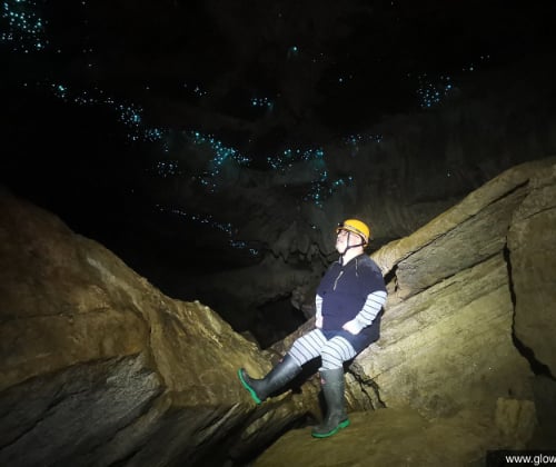 Visitor seated on cave rocks beneath glowworms, emphasizing the cave’s height and glowing ceiling.
