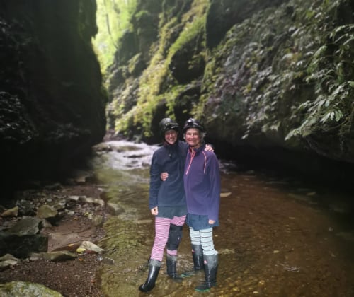 Two visitors standing in a shallow stream within a forested limestone gorge near the cave entrance.