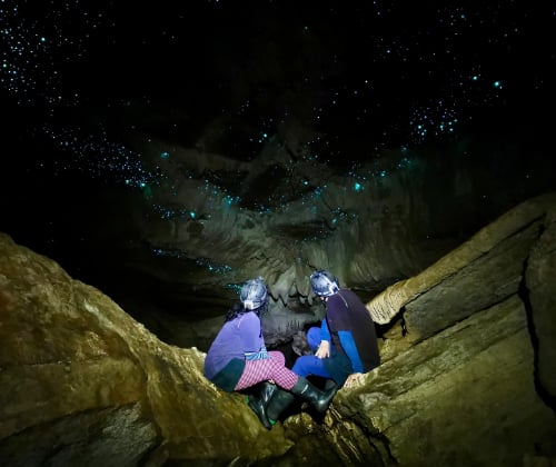 Two visitors sitting on cave rocks looking up at glowworms during a Down to Earth Glowworm Cave Tour in Waitomo.